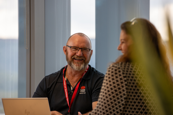 Person with beard and glasses seated at laptop facing other person with long hair, both smiling.
