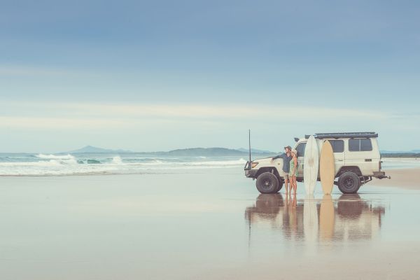 A white four wheel drive is parked on a beach. Two people stand beside it looking towards the ocean. Two surfboards - one white and one yellow - lean against the 4WD beside them