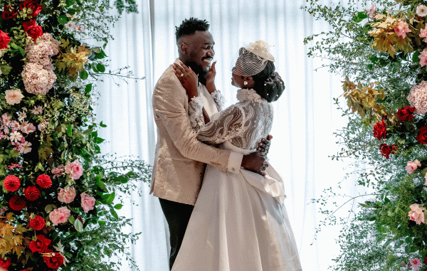 A bride wearing a white dress and groom in a beige suit jacket smile as they embrace at a floral altar.