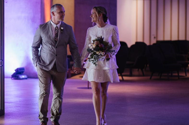 A bride and groom walk through a room lit by pink and purple linking